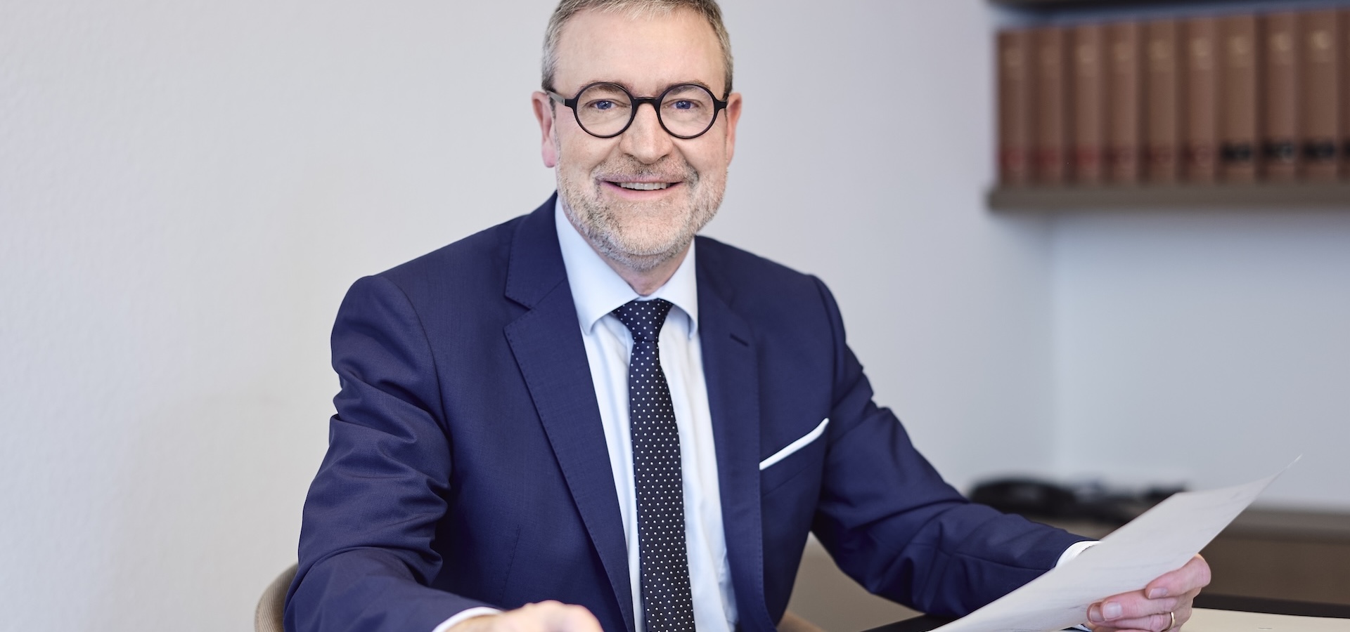 Dr. Matthias Breucker sitzt lächelnd am Schreibtisch, hält ein Dokument, dunkler Anzug, Bücherregal im Hintergrund