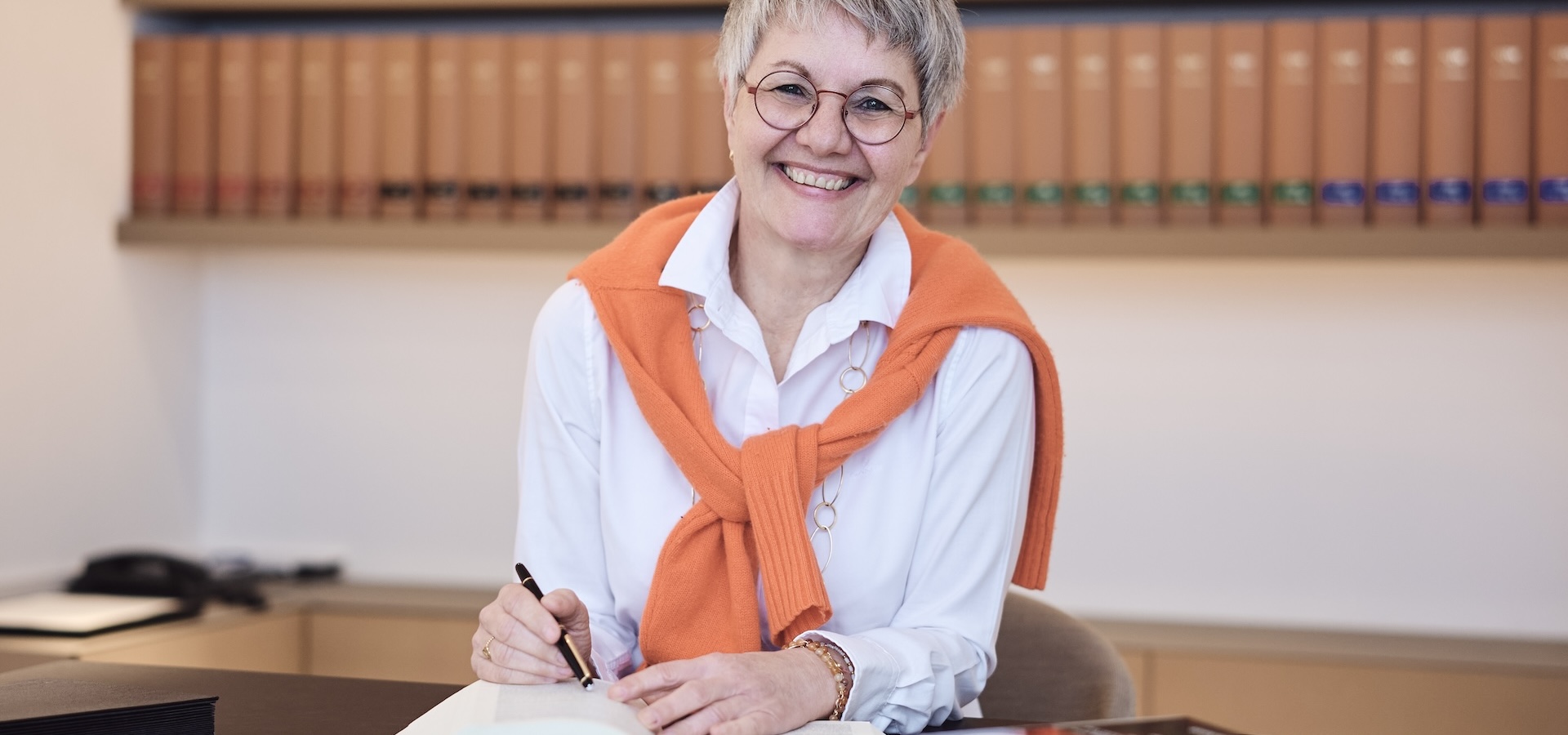 Anette Breucker lächelt am Schreibtisch, hält einen Stift, weiße Bluse, orangefarbene Strickjacke über den Schultern